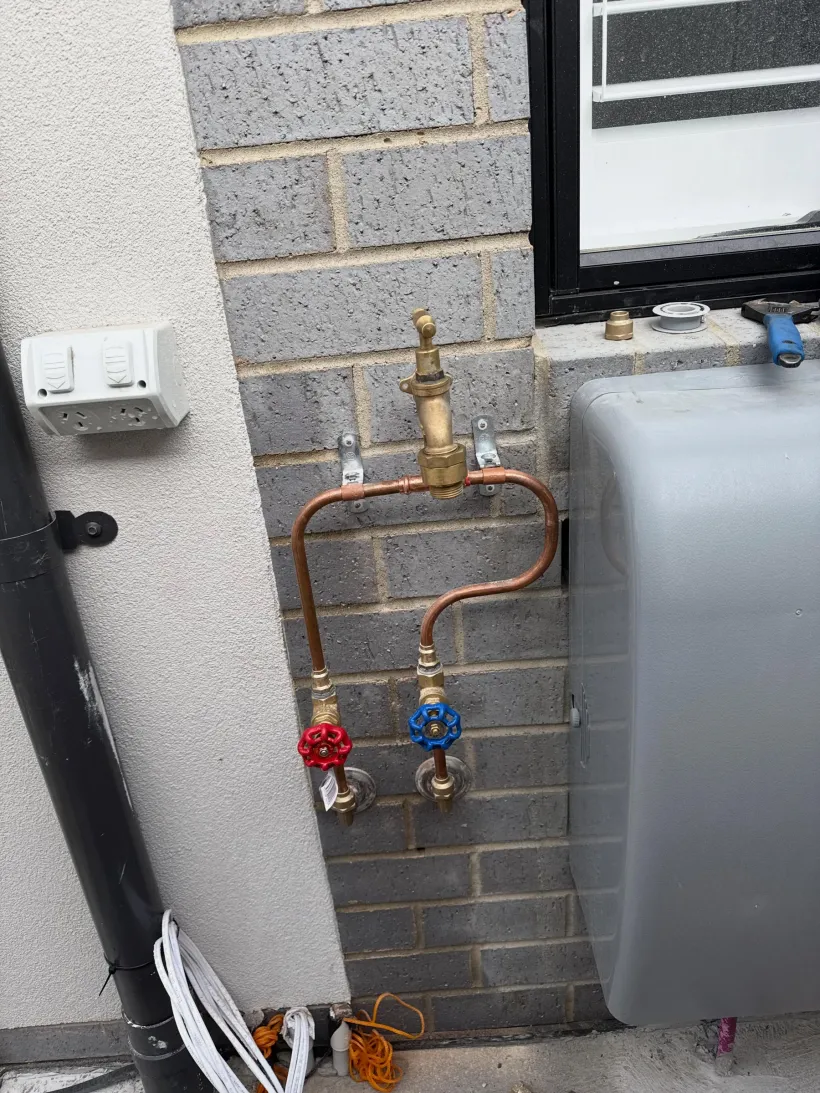 Copper pipework with a brass tempering valve above red and blue isolation valves, mounted on a brick wall beside a hot water unit