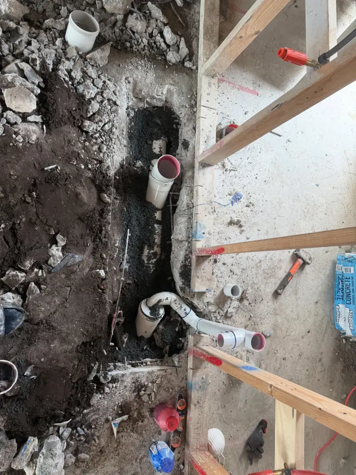 Excavated drainage rough-in with white PVC pipe and junctions laid on a gravel bed beside timber formwork