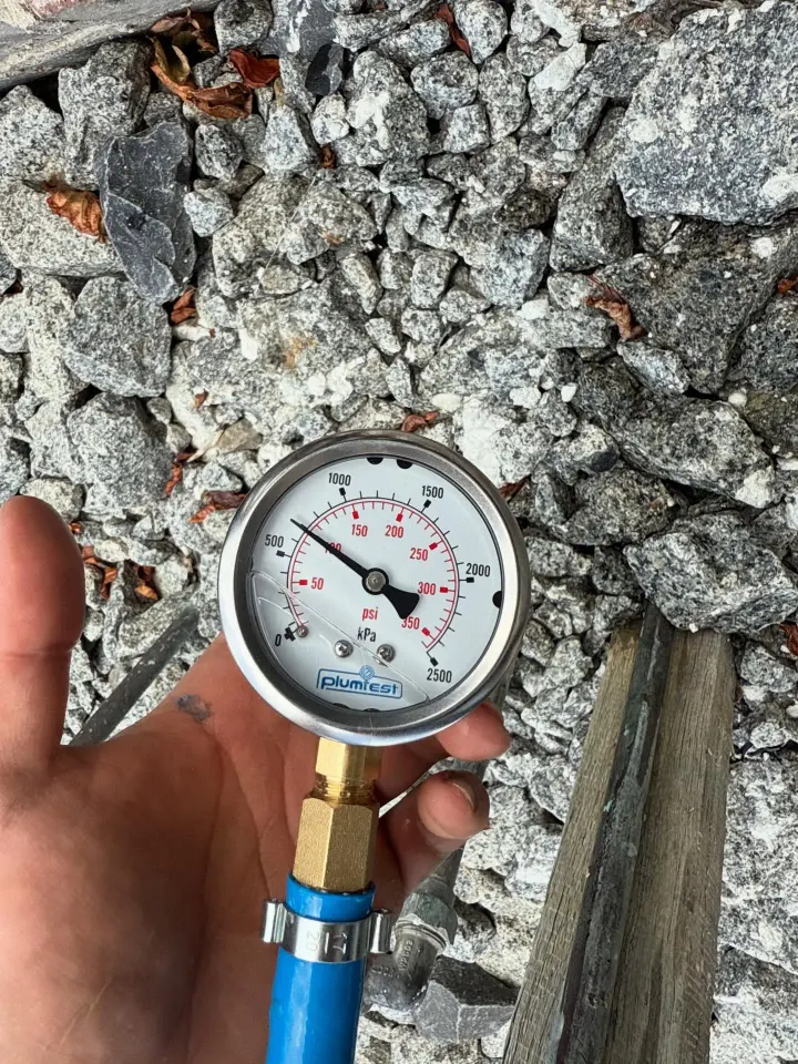 Plumber holding a pressure test gauge connected to a blue test hose, reading just under 500 kPa