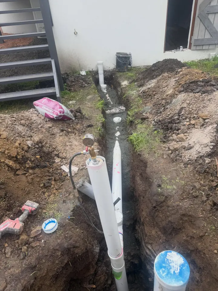 Long open trench with sewer pipework laid on a gravel bed alongside a rendered wall